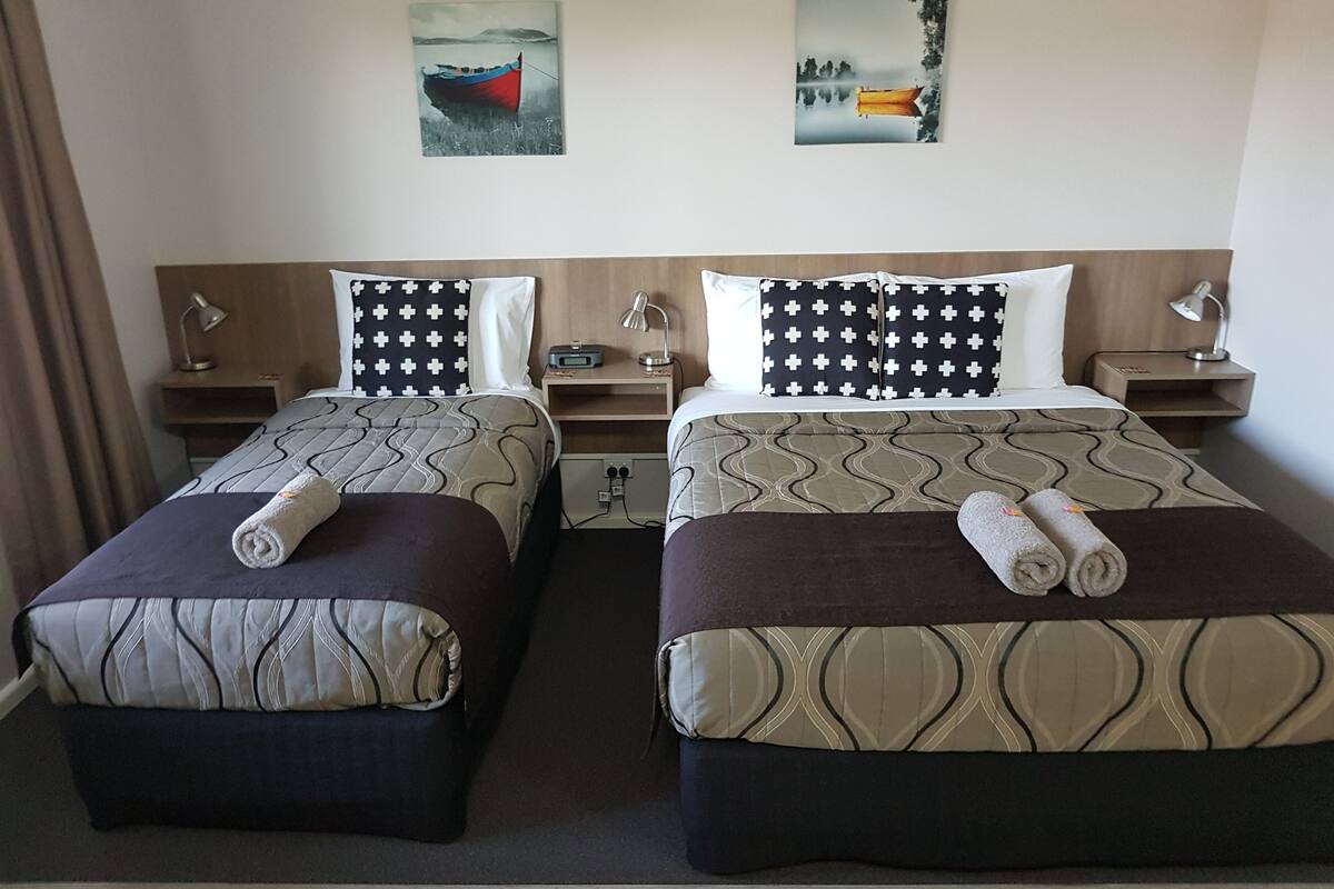 A hotel room with two beds, each with a patterned bedspread, pillows with a cross design, bedside tables with lamps, and artwork on the walls.