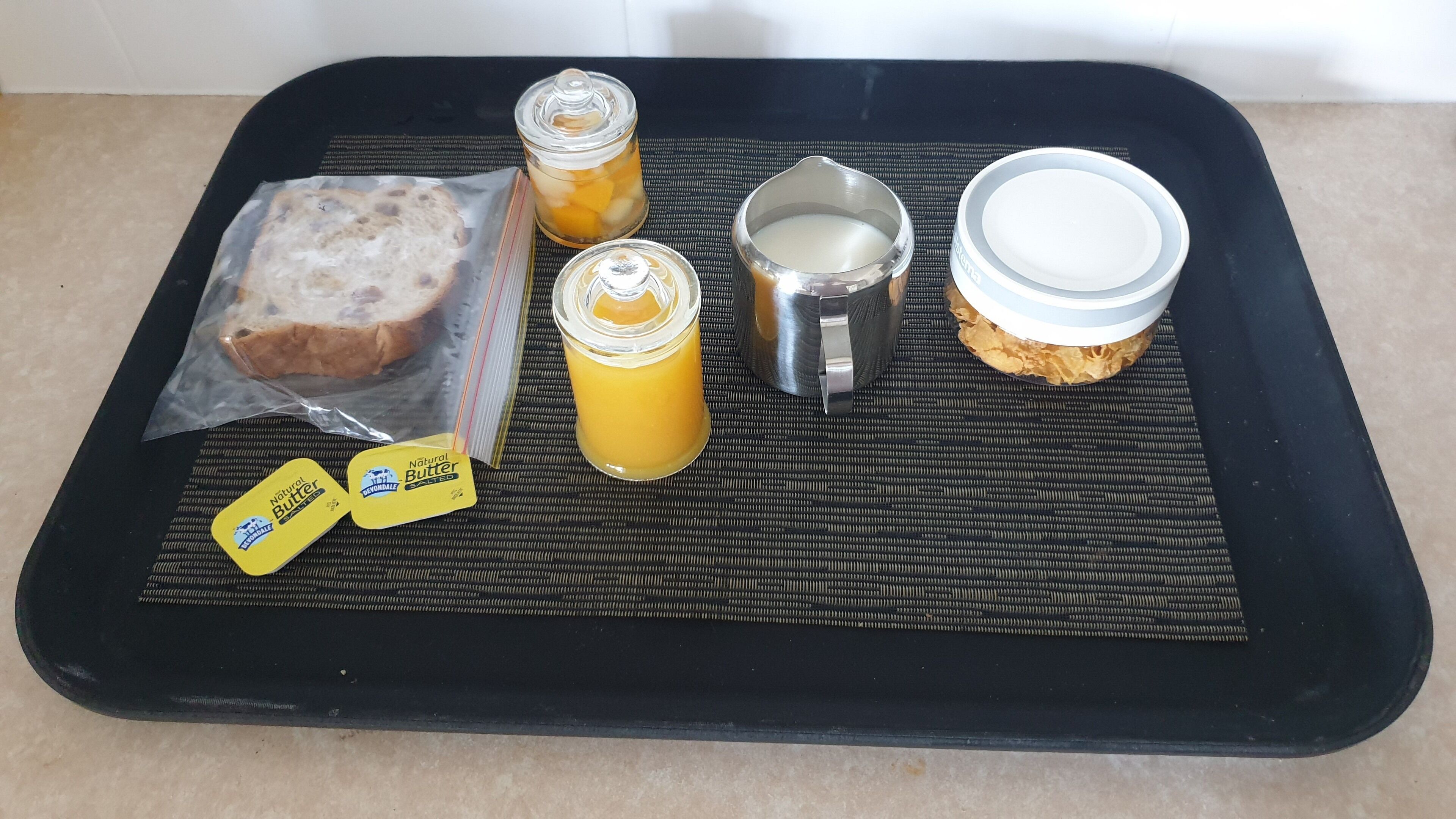 Ein Tablett mit Brot, zwei Buttergläsern, einem Metallbehälter und einem Orangensaftglas.