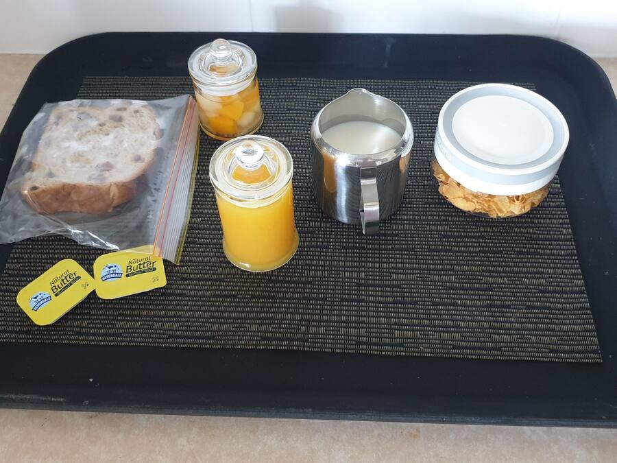 Una bandeja con hogaza de pan, dos recipientes de mantequilla, un contenedor metálico y un frasco de jugo de naranja.