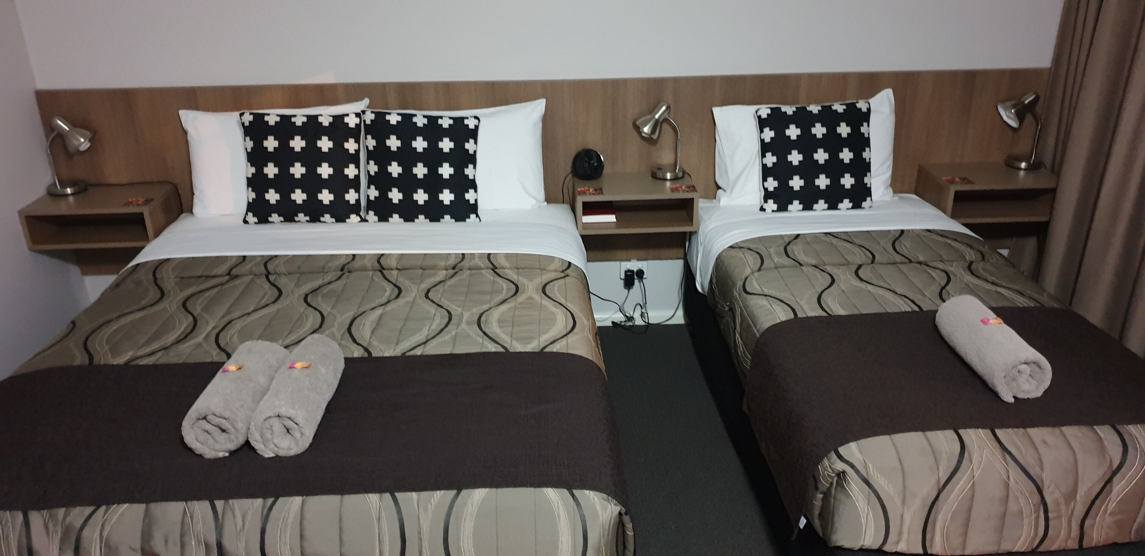 A hotel room with two beds, each with patterned bedding and pillows, bedside lamps, and rolled towels on the beds.