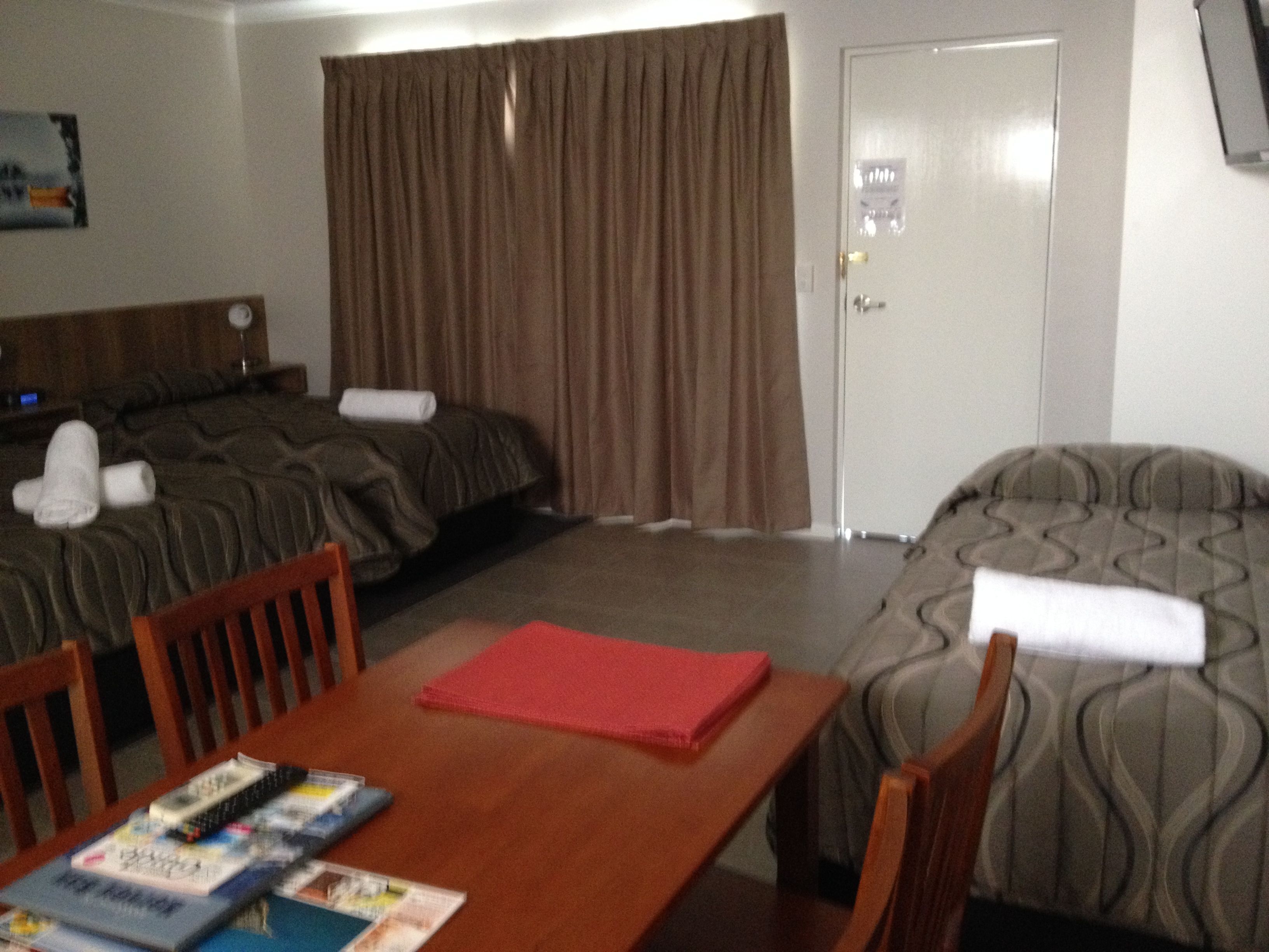 A hotel room with two beds, a wooden table with magazines, and a door with a light switch.