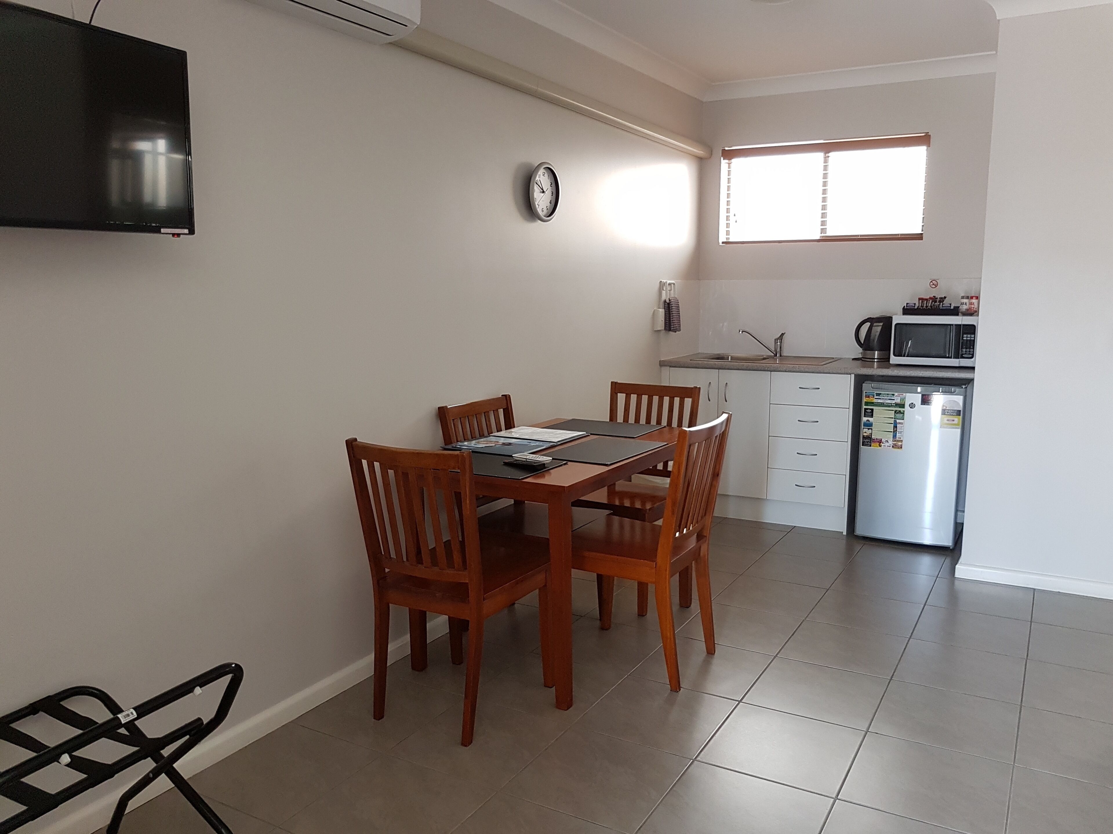 A dining area with a wooden table and chairs, a clock on the wall, a kitchen with a refrigerator, microwave, and sink.