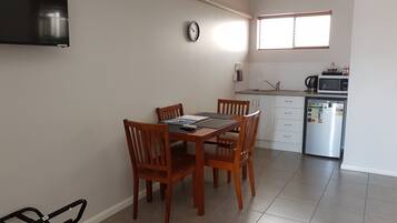 A dining area with a wooden table and chairs, a clock on the wall, a kitchen with a refrigerator, microwave, and sink.
