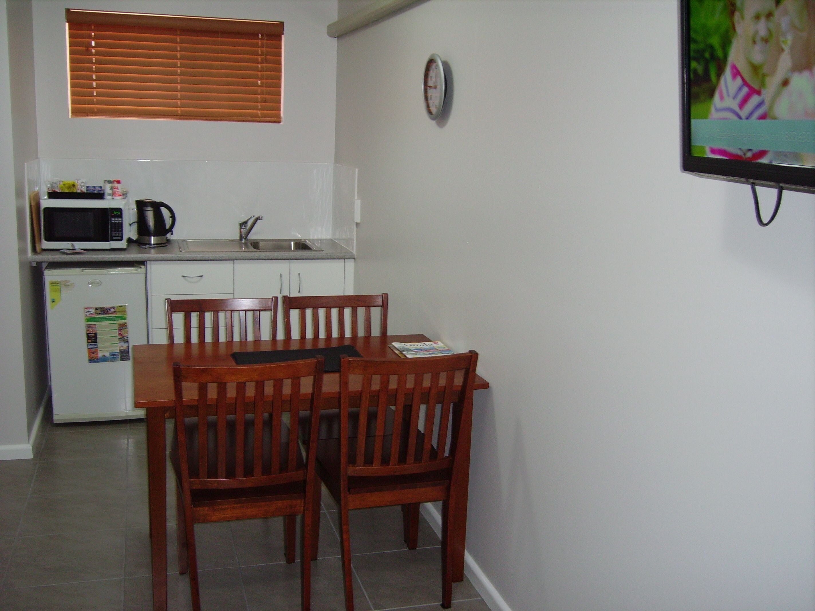A compact kitchen with a microwave, kettle, and refrigerator. There is a dining table with chairs and a clock on the wall.
