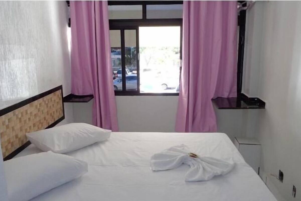 A hotel room with a bed, white pillows, a window with pink curtains, and a view of the outside.