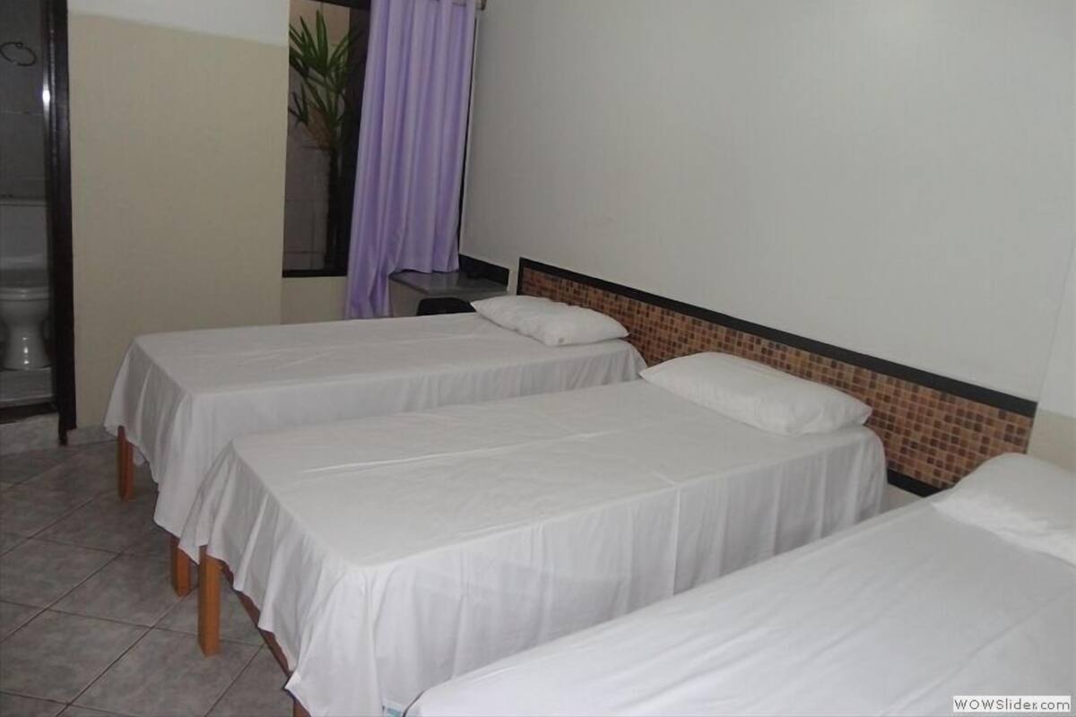 A room with three single beds, a tiled floor, a window with a curtain, and a small table.