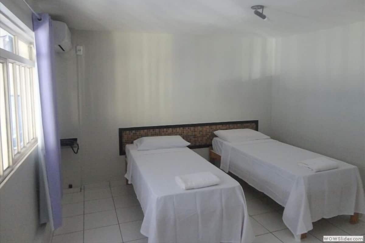 A room with two single beds, a window with blinds, and a ceiling light.