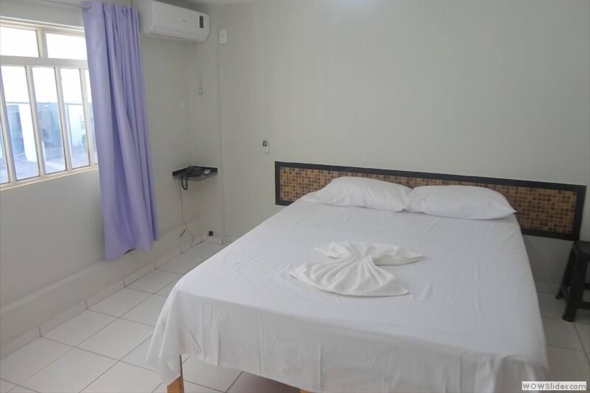 A hotel room with a bed, a window with curtains, an air conditioning unit, and a tiled floor.