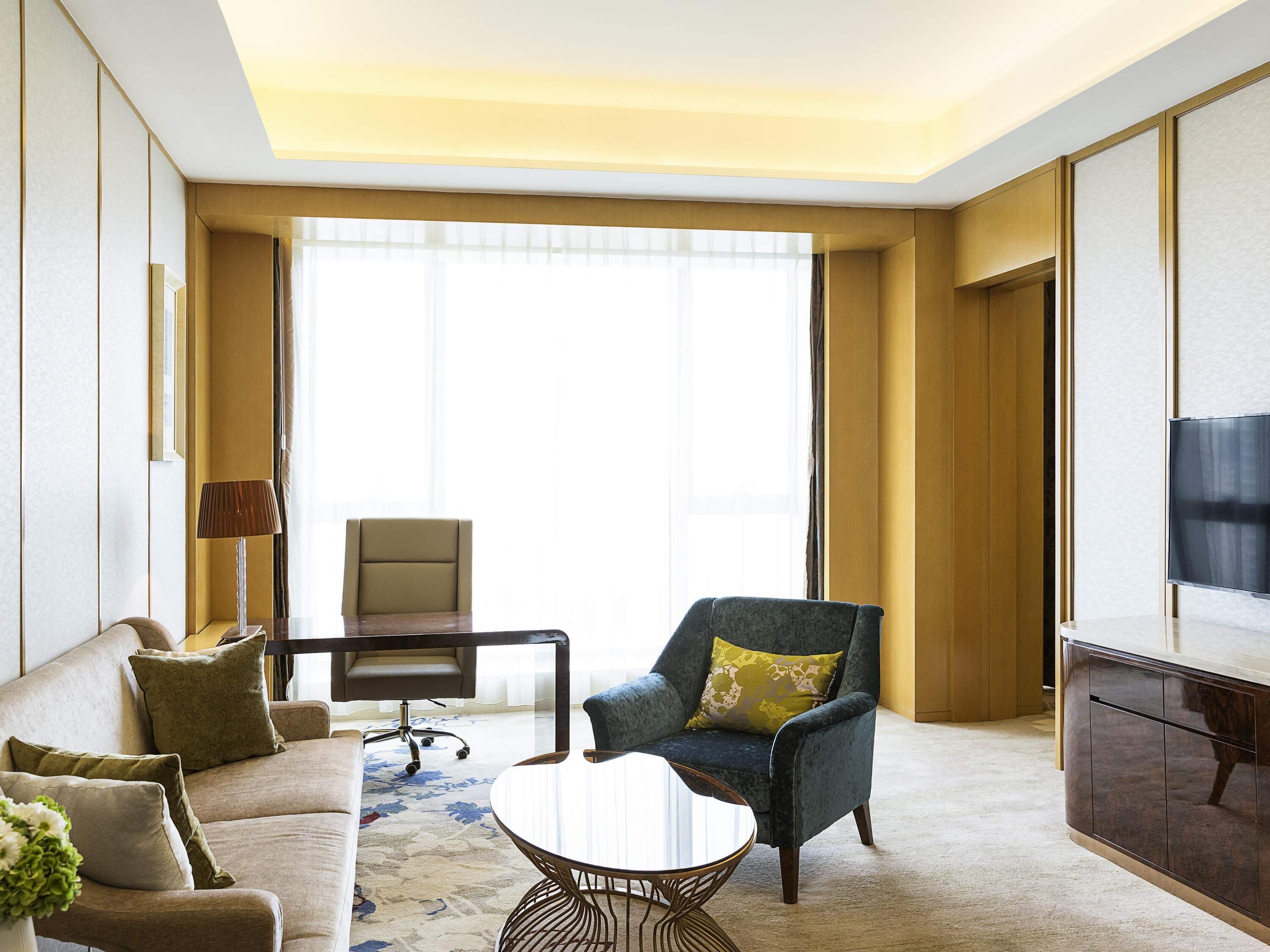 A modern hotel room with a sofa, armchair, coffee table, desk, and TV.