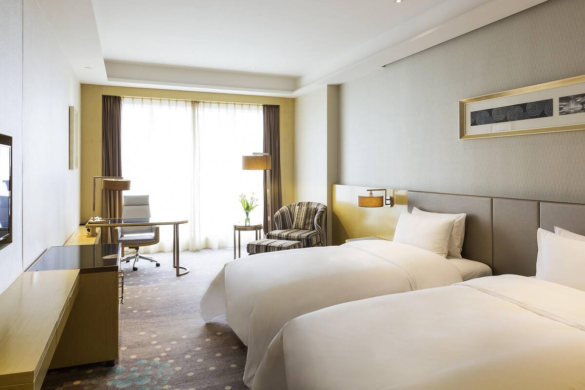 A hotel room with two beds, a desk, a chair, a lamp, and a framed picture on the wall.