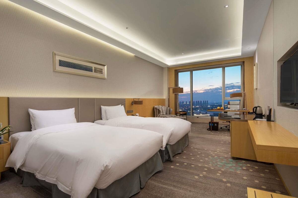 A hotel room with two beds, a desk, a chair, a large window with a city view, and a mounted air conditioner.