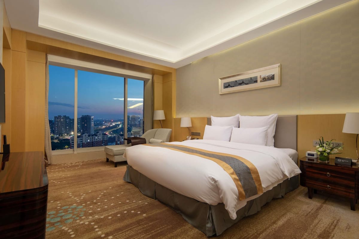 A hotel room with a large bed, a desk, a chair, a nightstand, and a view of the city at dusk.