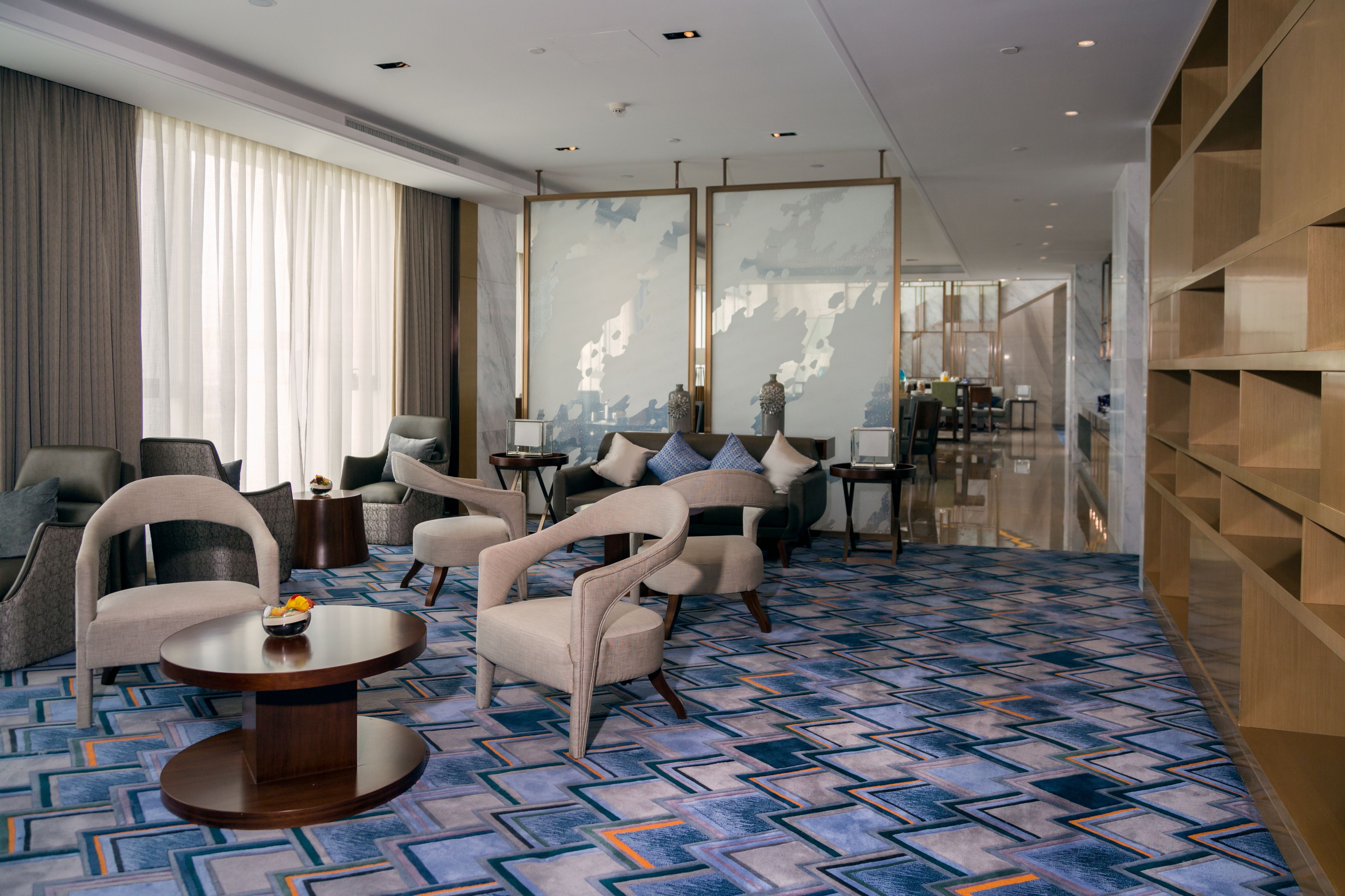 A modern hotel lobby with a geometric patterned carpet, contemporary furniture, and a large mirror reflecting the room.