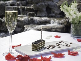 A dessert plate with a layered cake and chocolate sauce, a glass of champagne, and red rose petals on a white tablecloth.