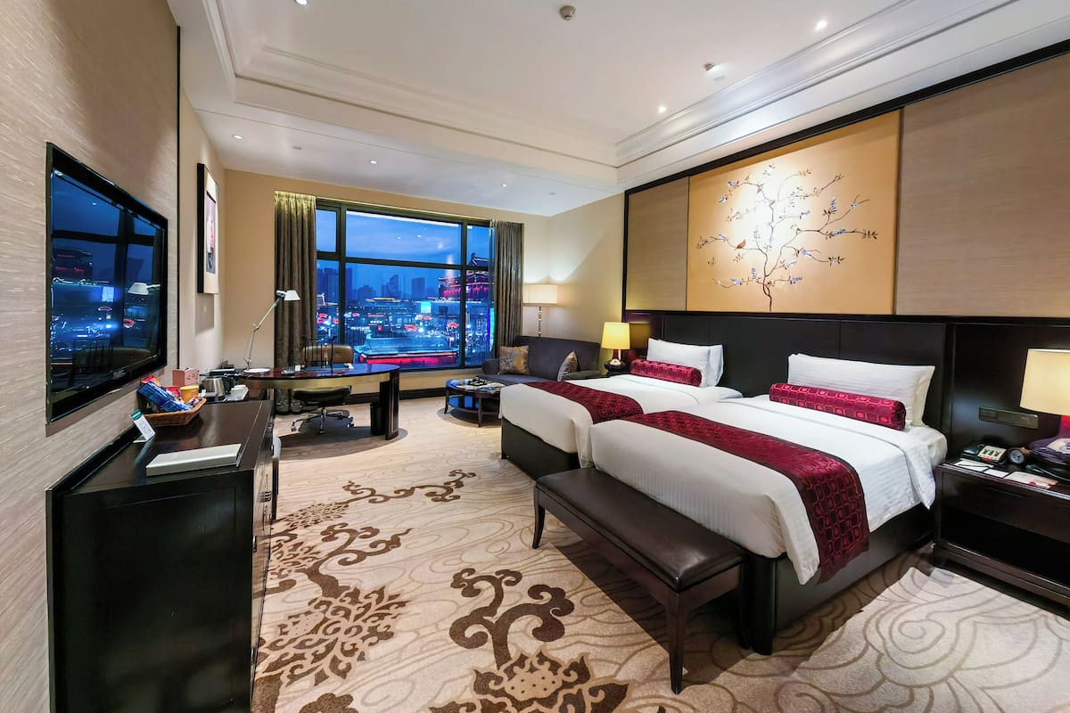 A hotel room with two beds, a desk, a chair, a TV, and a large window with a city view.