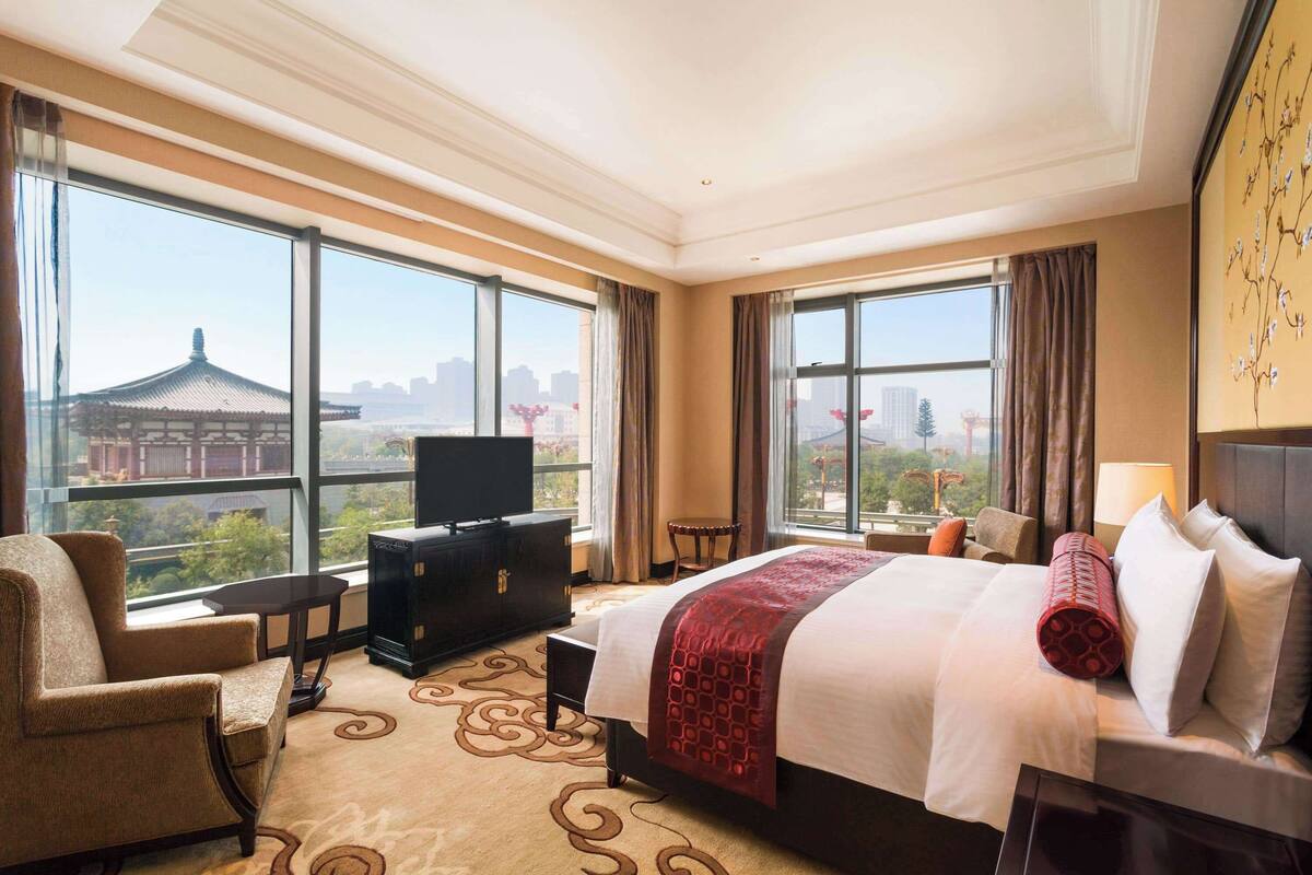 A hotel room with a large bed, a TV, a chair, and a view of a cityscape.
