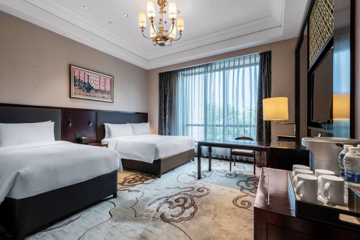 A hotel room with two beds, a desk, a chair, a lamp, a coffee station, and a large window with curtains.
