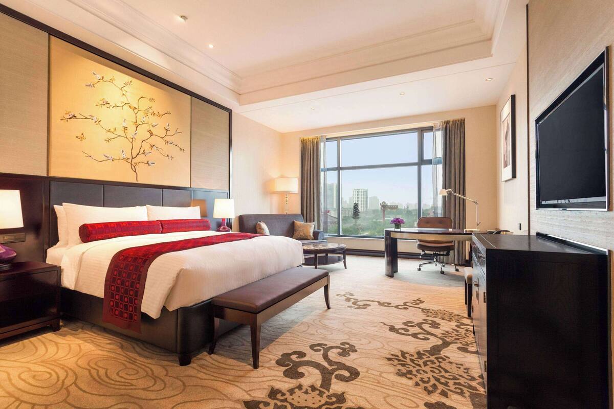 A hotel room with a large bed, a desk, a chair, a TV, and a window with a city view.