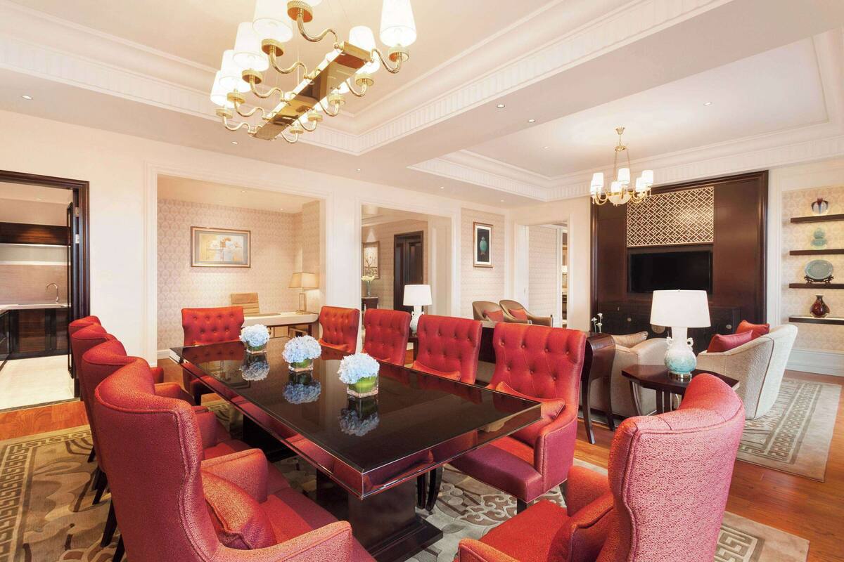 A spacious room with a large rectangular table surrounded by red upholstered chairs, a chandelier, and a television.