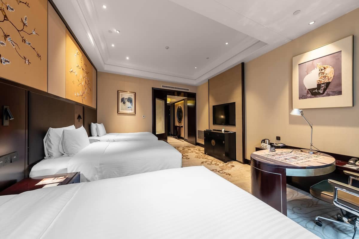 A hotel room with two beds, a desk with a lamp, a TV, and artwork on the walls.