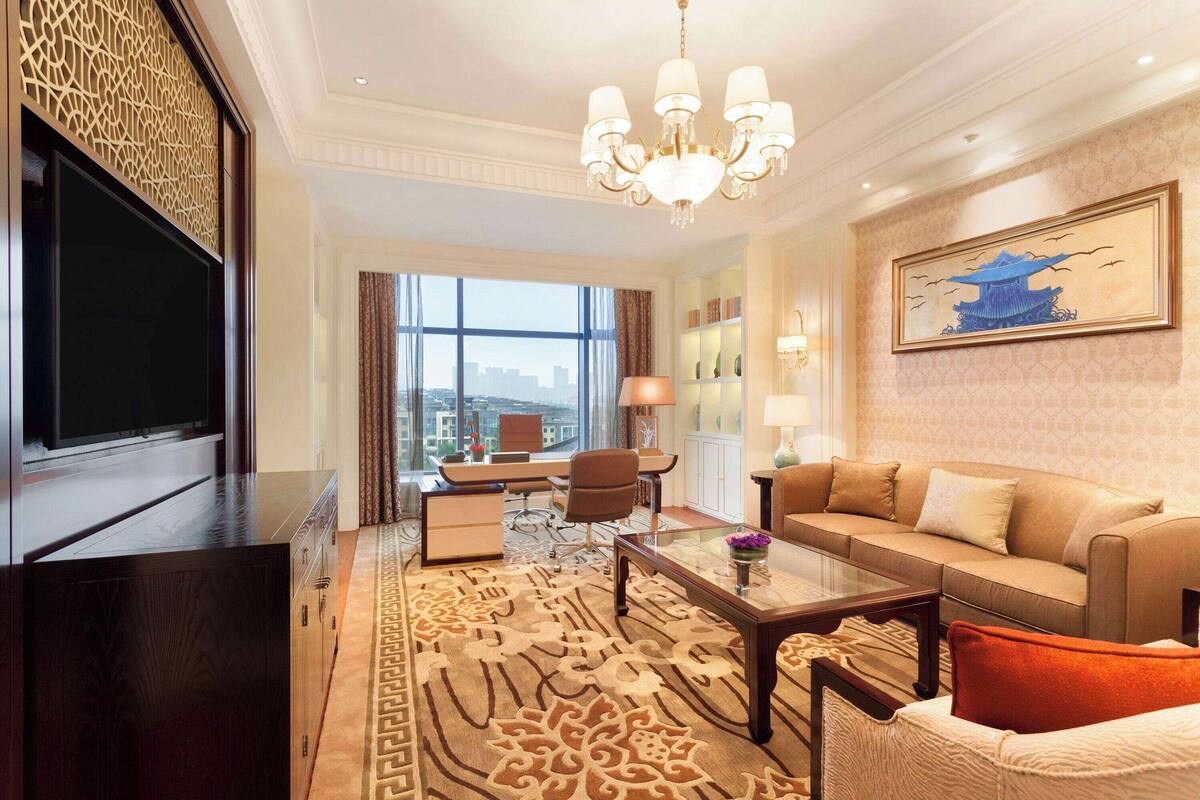 A hotel room with a large TV, a sofa, a coffee table, and a desk with a chair. There is a large window with a view of the city.