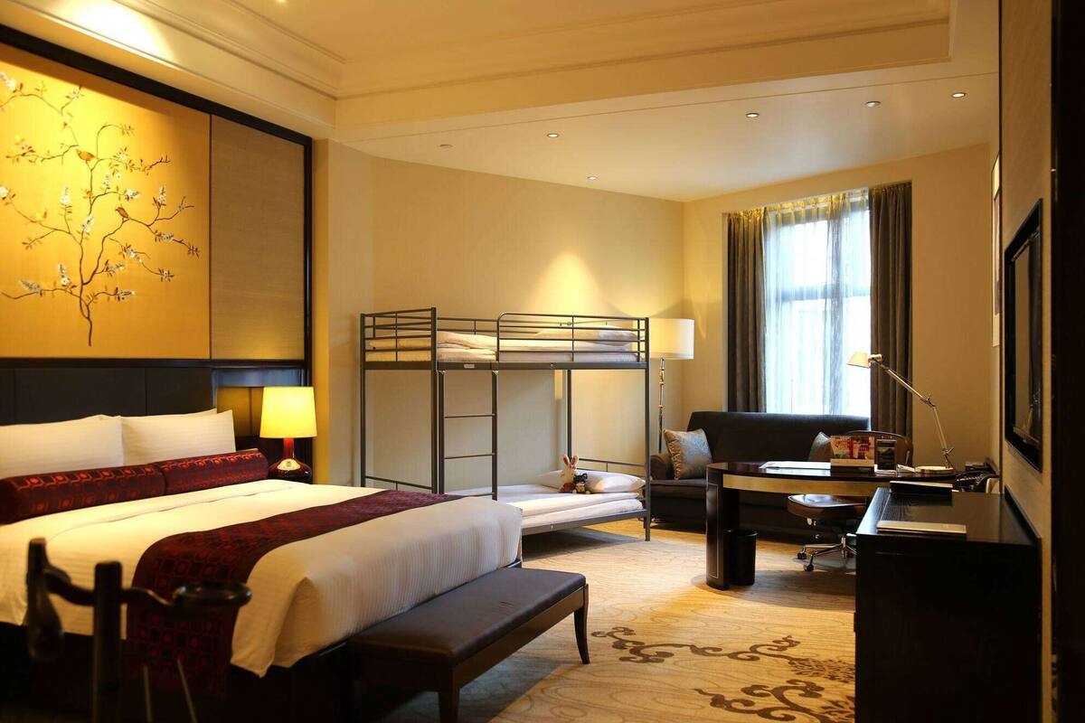 A hotel room with a bunk bed, a desk with a chair, a lamp, a large window with curtains, and a decorative wall piece.