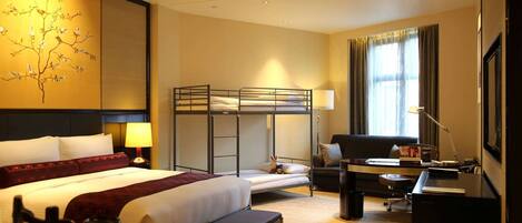 A hotel room with a bunk bed, a desk with a chair, a lamp, a large window with curtains, and a decorative wall piece.