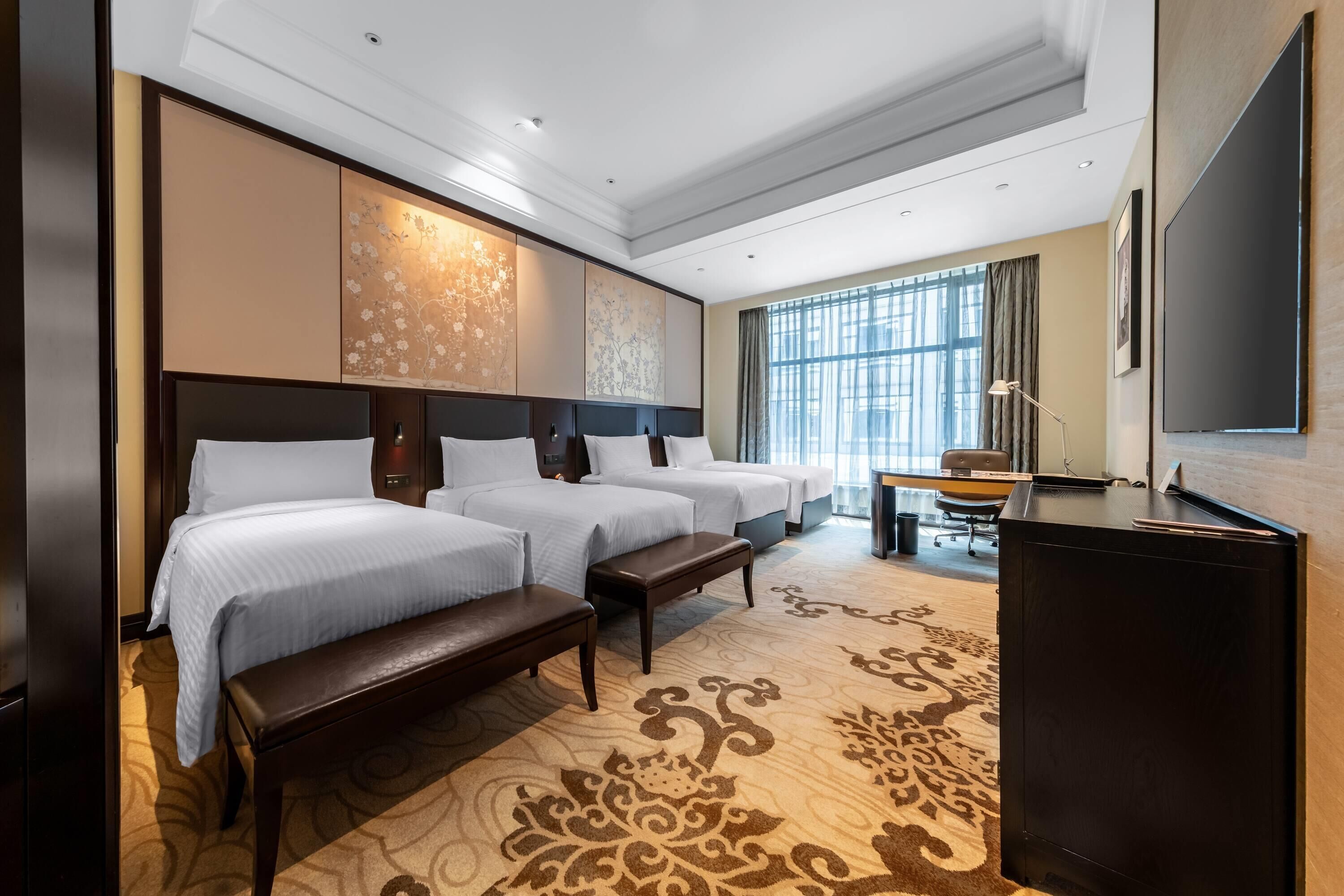 A hotel room with three beds, a desk, a television, and large windows.