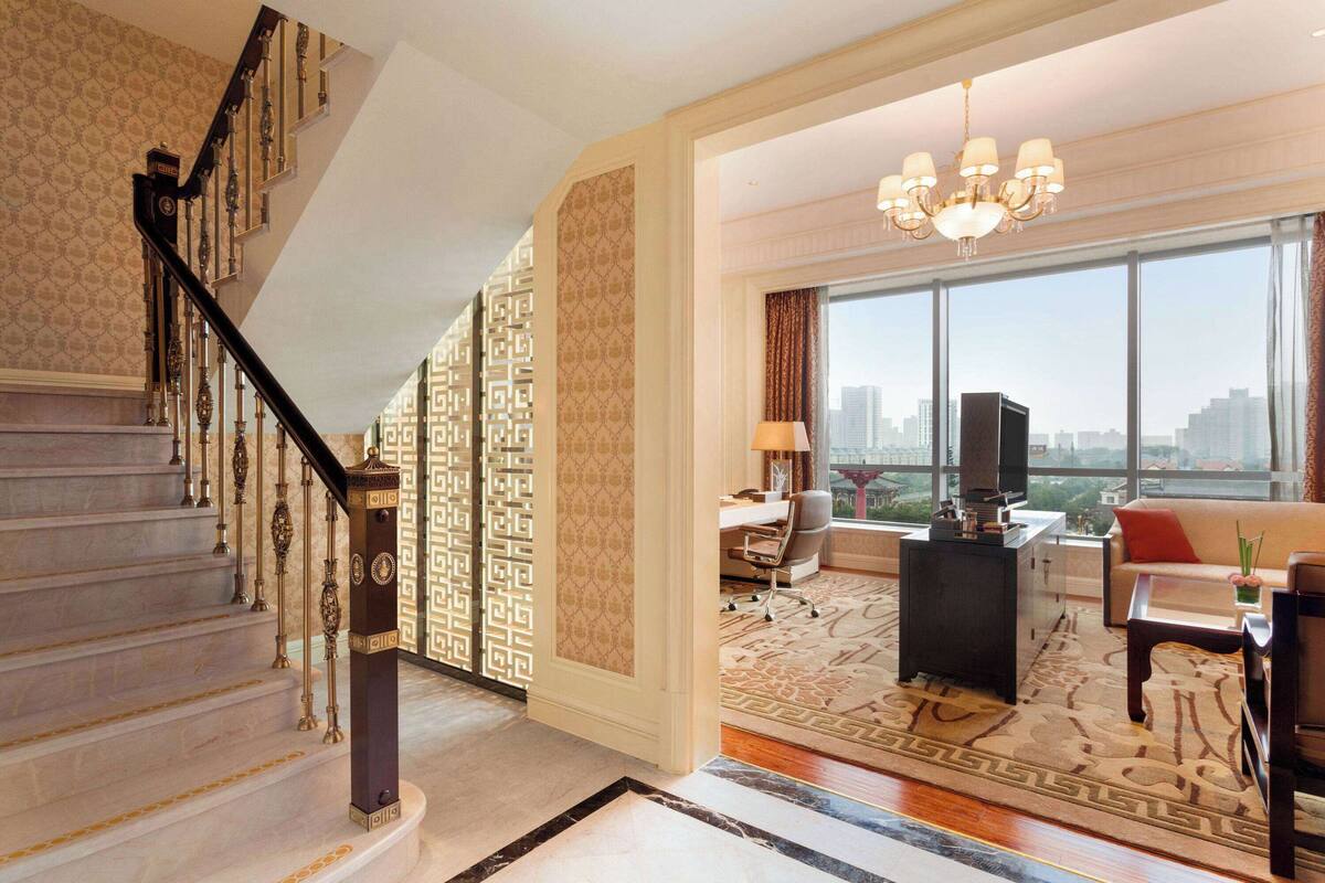 A spacious living area with a staircase, a large window with a city view, a chandelier, and a sofa.