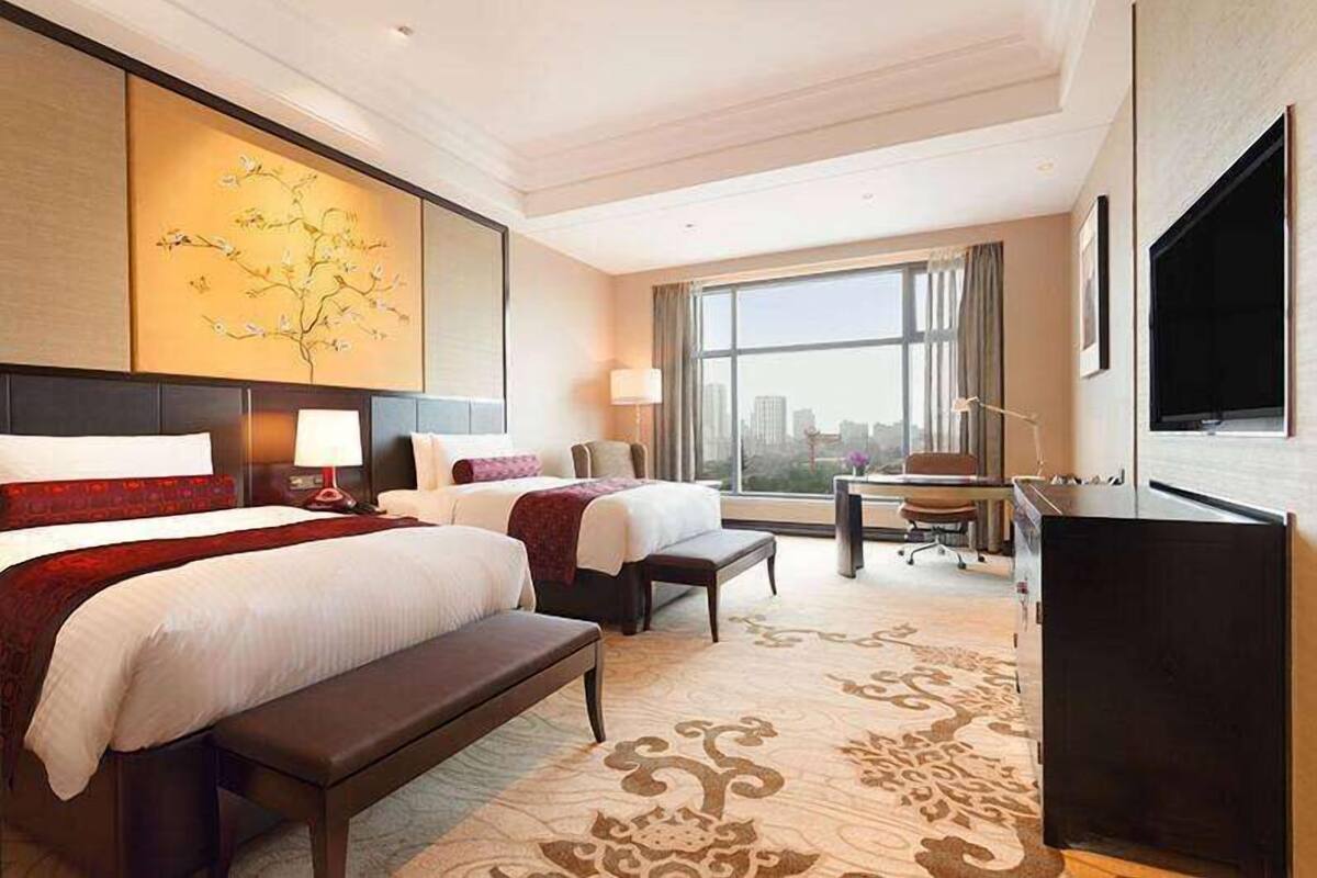 A hotel room with two beds, a desk, a chair, a large window with a city view, and a flat-screen TV.