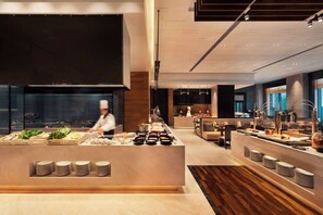 A modern buffet setup with a variety of food items and a chef preparing food in the background.