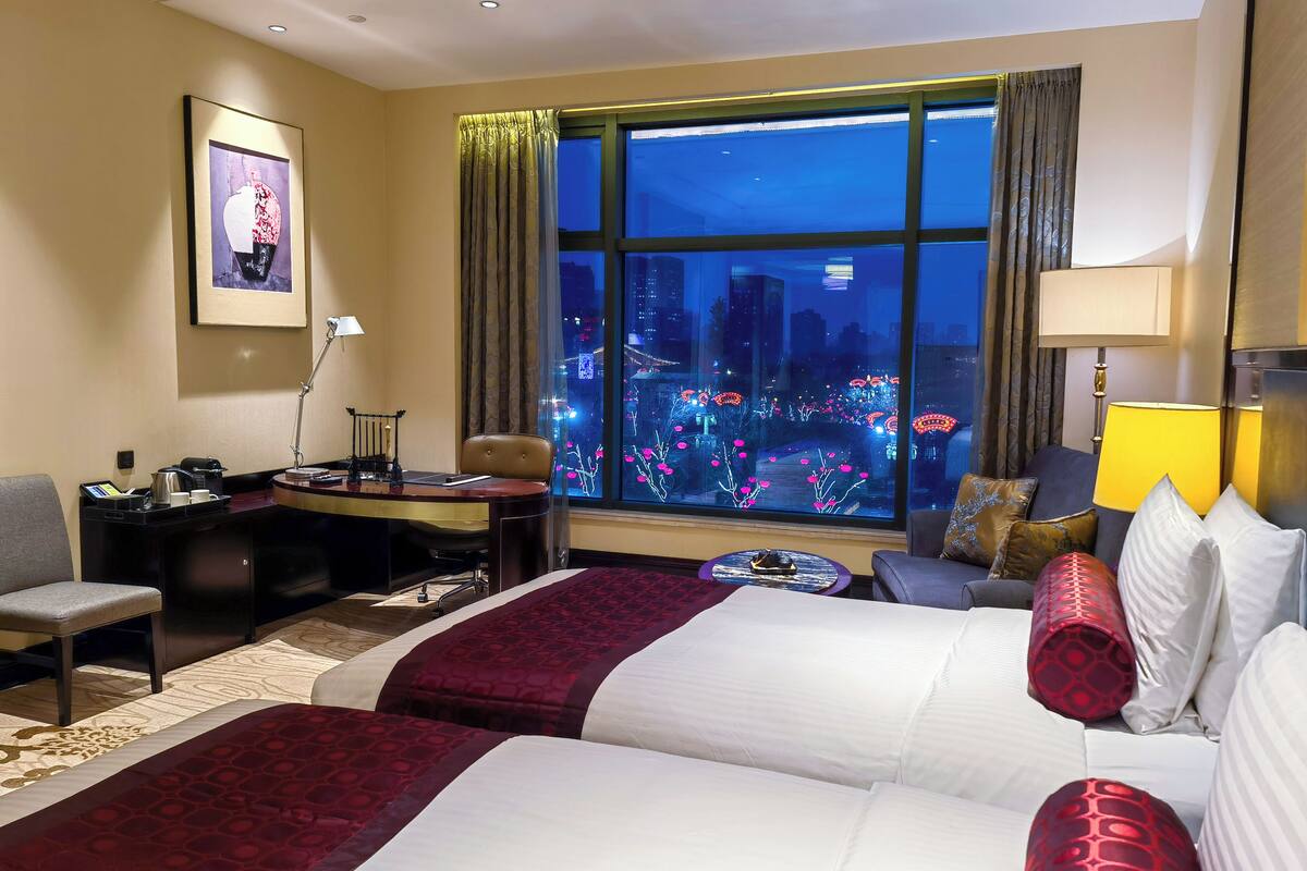 A hotel room with two beds, a desk, a chair, a small table, and a large window with a city view.