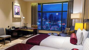 Habitación de hotel con dos camas, un escritorio, una silla, una mesita pequeña y un ventanal grande con vistas a la ciudad.