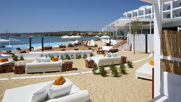 A beachside lounge area with white sofas, sun loungers, and umbrellas, overlooking the sea and a modern building with balconies. - Nikki Beach Resort & Spa Porto Heli (Ermionida)