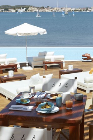 A dining area with a table set for breakfast, overlooking a body of water with sailboats. - Aliis Resort Porto Heli (Ermionida)