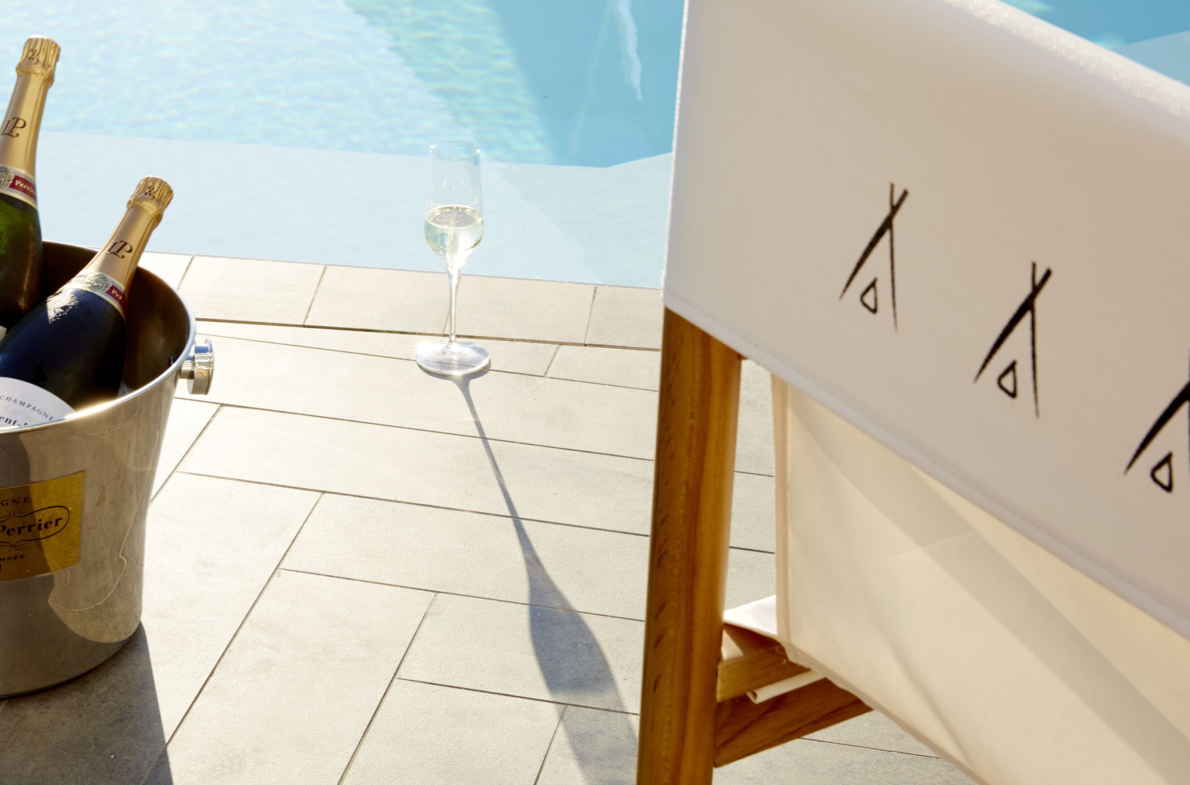 A champagne bucket with bottles and a glass on a tiled floor, with a pool in the background.