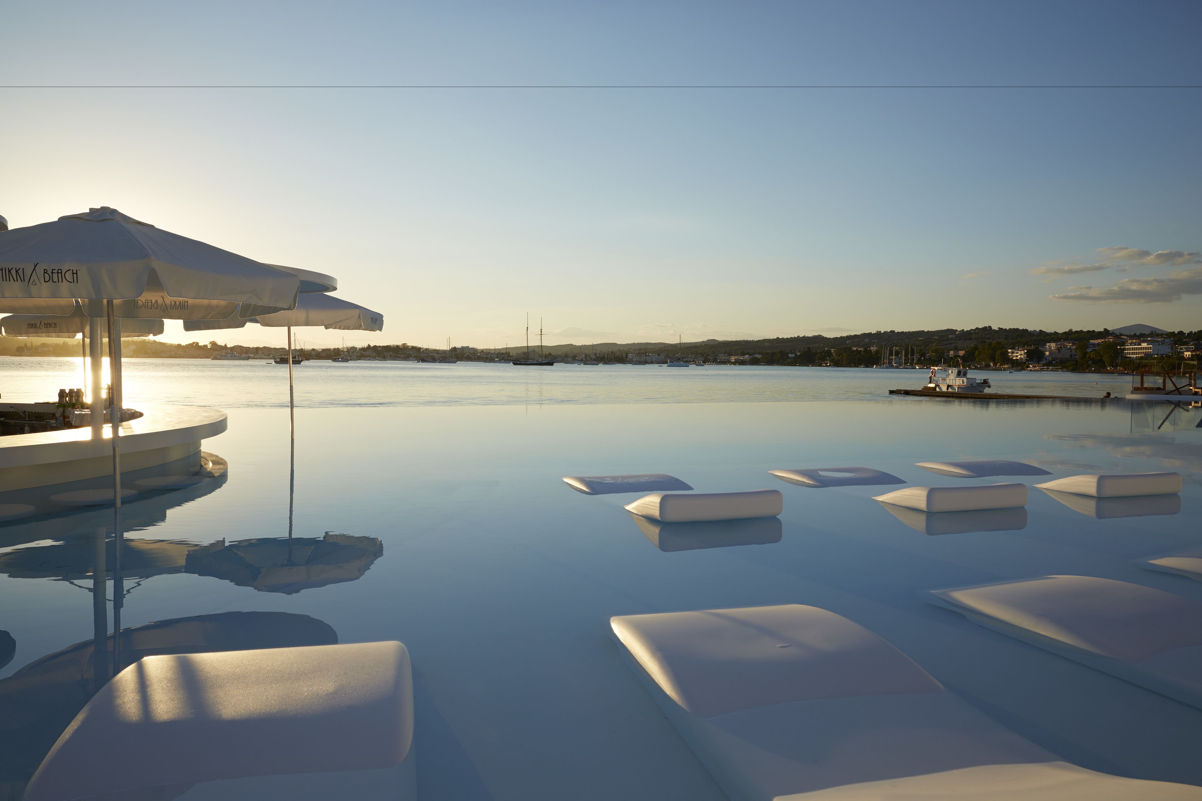 A serene infinity pool with floating lounge chairs and a sun umbrella, overlooking a calm body of water with distant buildings.