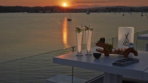 A table with three tall glasses of a clear beverage, a candle, and a small bowl, set against a backdrop of a sunset over a calm body of water with boats. - Nikki Beach Resort & Spa Porto Heli (Ermionida)