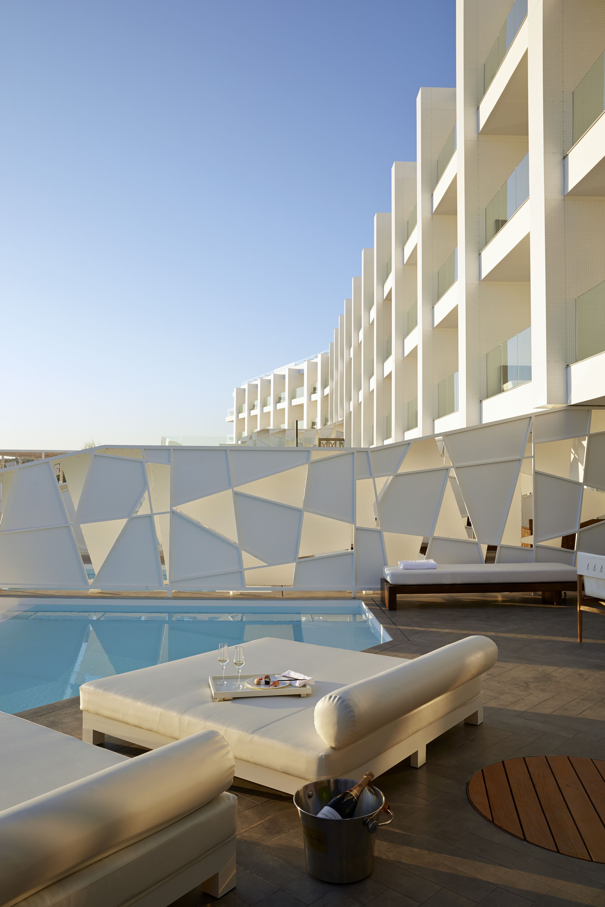 Un espace aménagé au bord de la piscine, avec des chaises longues blanches, une piscine et un bâtiment moderne aux motifs géométriques en arrière-plan.