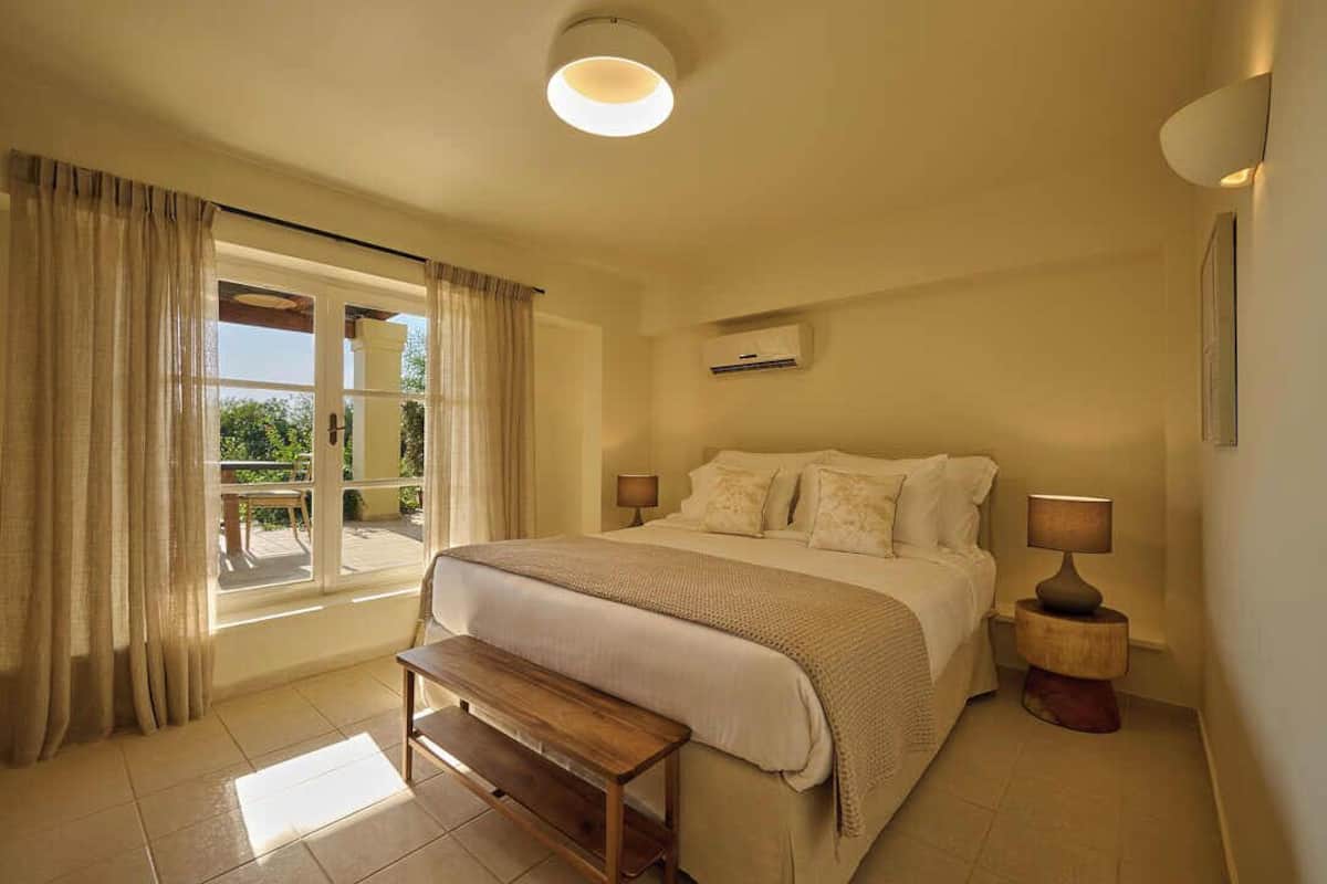 Ein Schlafzimmer mit einem Bett, Nachttischlampen, einer Bank, einem Fenster mit Vorhängen und einer Klimaanlage.
