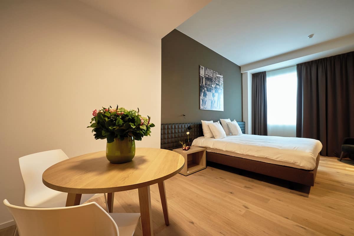 A modern hotel room with a wooden floor, a round dining table with chairs, a bed with a headboard, a nightstand, and a wall-mounted picture.