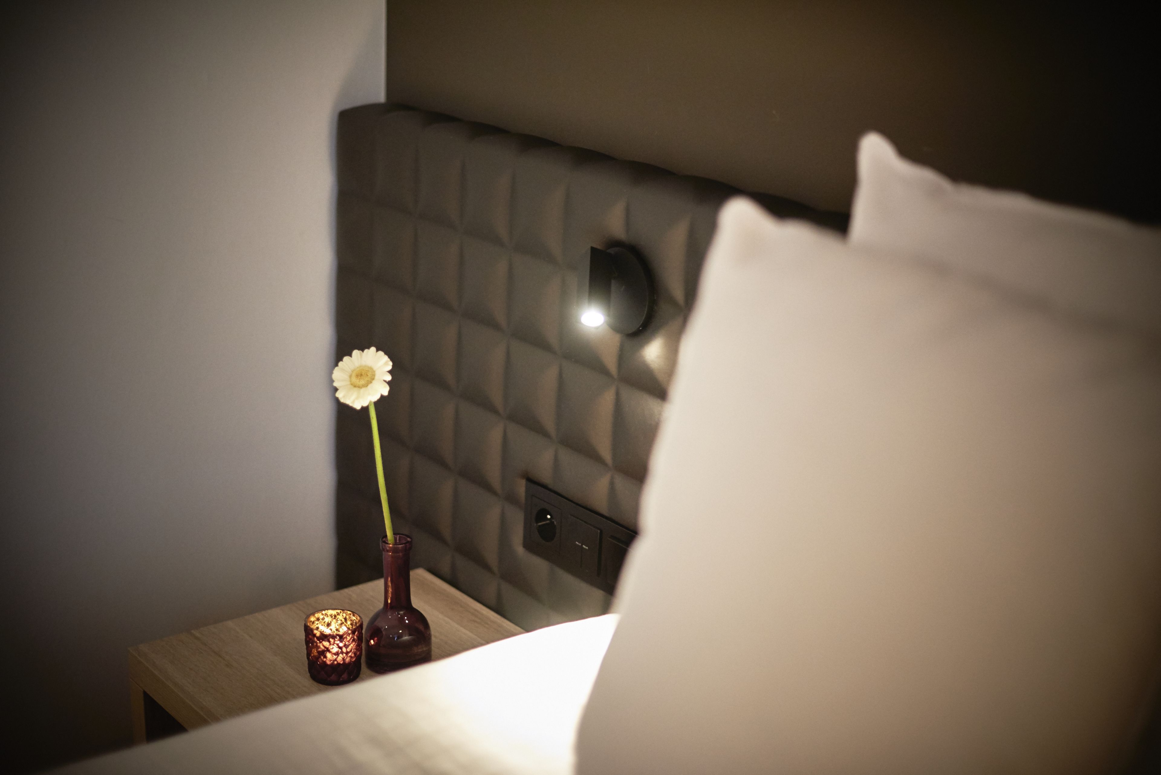 A neatly made bed with a white pillow, a small vase with a single white flower, and a lit candle on a bedside table.