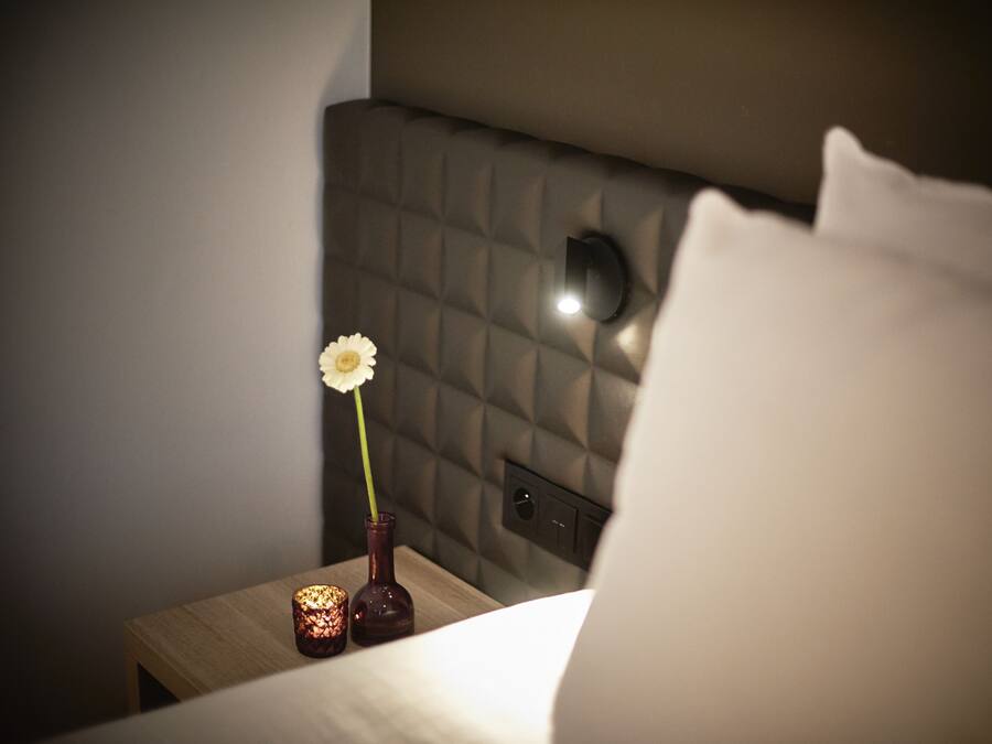 Ein ordentlich bezogenes Bett mit einem weißen Kissen, ein kleiner Strauß mit einer einzelnen weißen Blume und eine brennende Kerze auf einem Nachttisch.