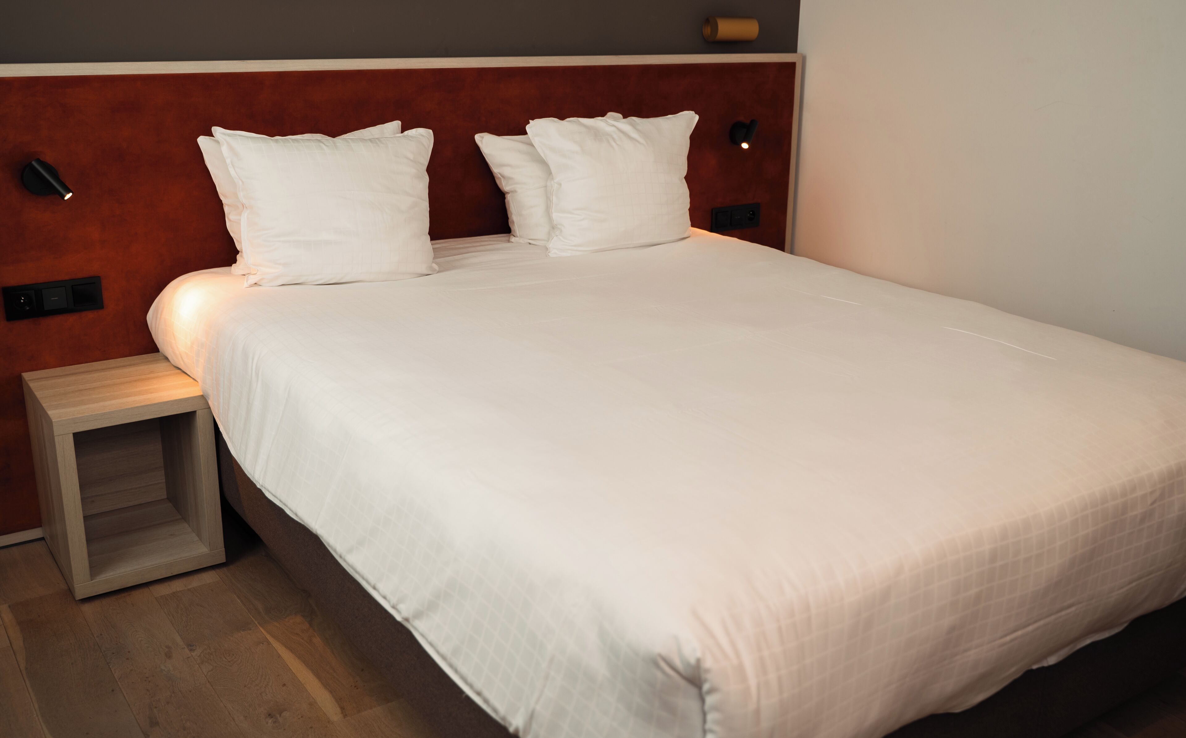 A neatly made bed with white linens and pillows, a wooden nightstand, and a wall-mounted light fixture.