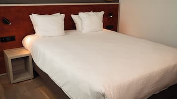 A neatly made bed with white linens and pillows, a wooden nightstand, and a wall-mounted light fixture.