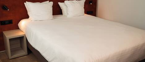 A neatly made bed with white linens and pillows, a wooden nightstand, and a wall-mounted light fixture.