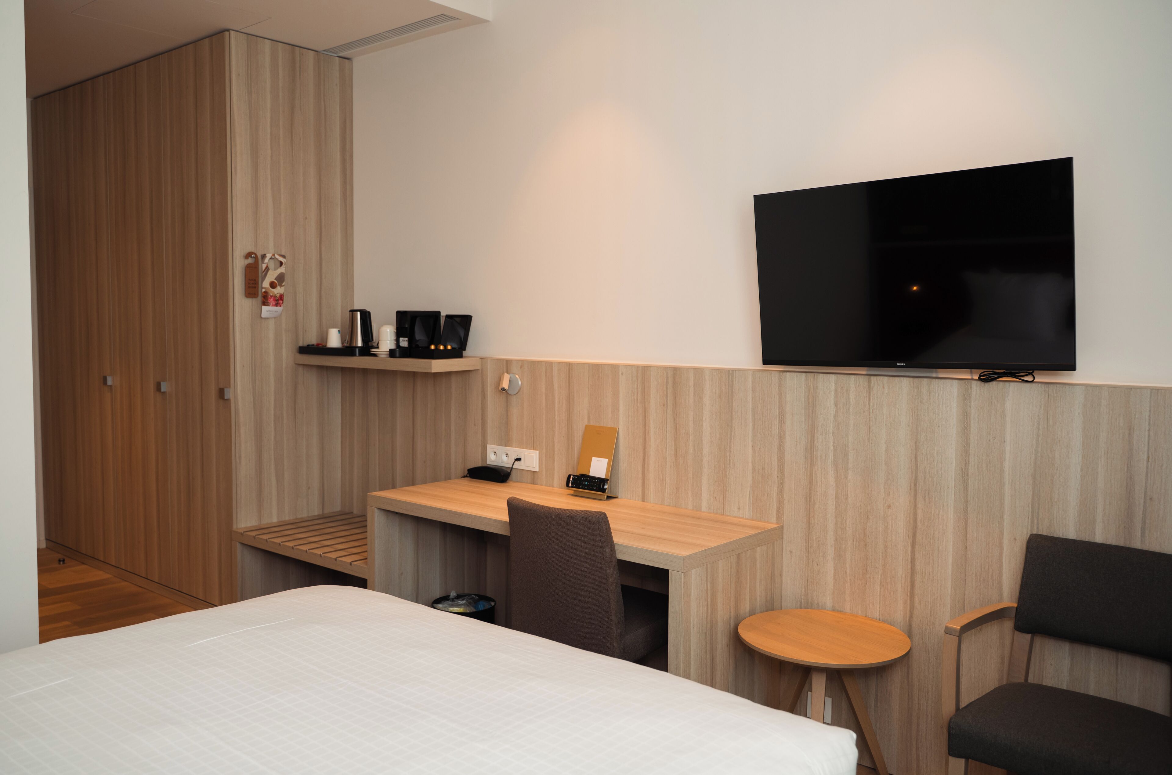 A hotel room with a bed, a desk with a chair, a TV mounted on the wall, and a small table.
