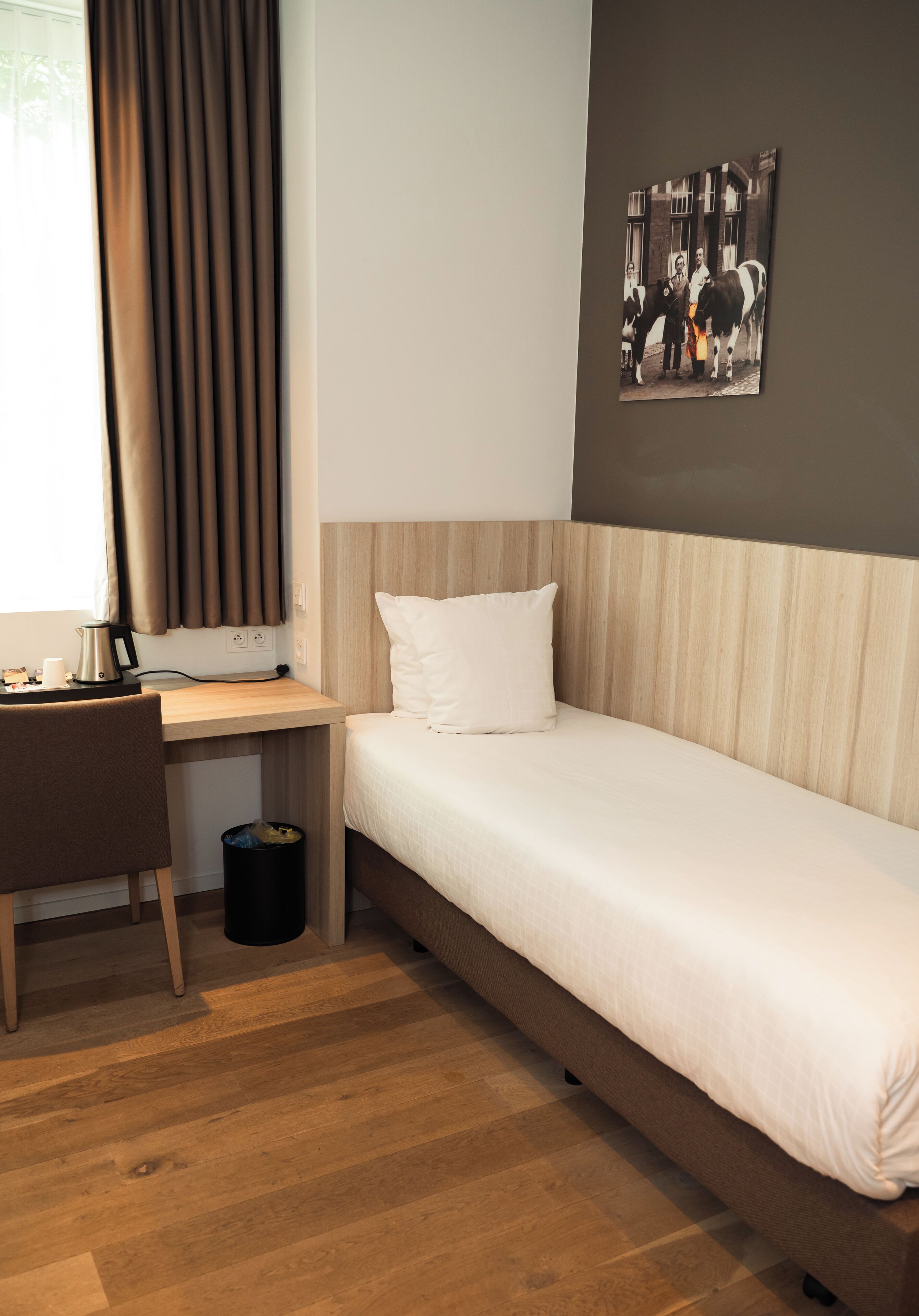 Een moderne hotelkamer met een houten bureau, een enkel bed, een aan de muur bevestigd schilderij en een raam met gordijnen.