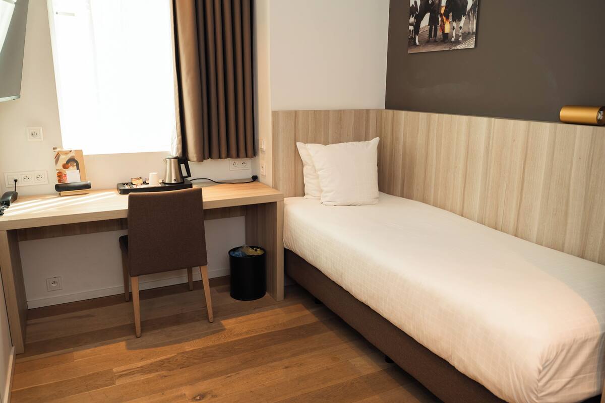 A compact hotel room with a single bed, a desk with a chair, a small trash bin, and a window with curtains.