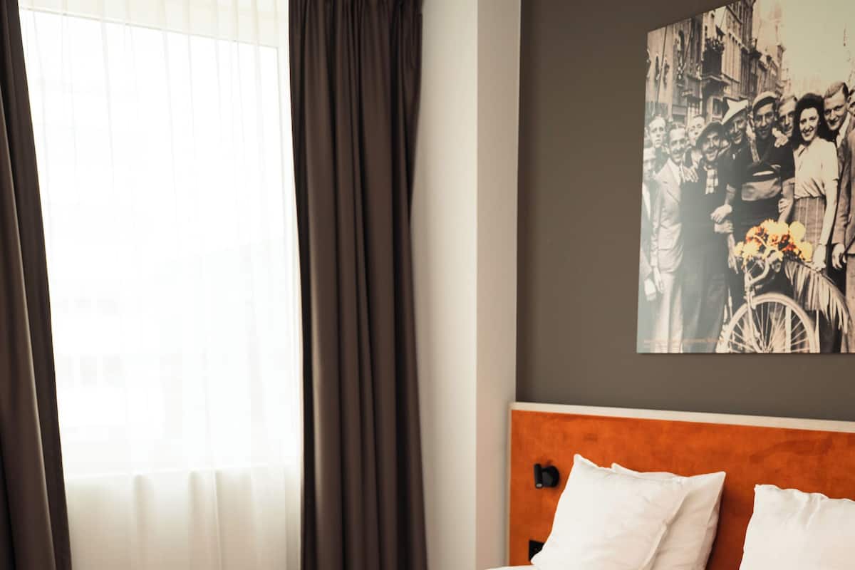A neatly made bed with white linens in a room with a wooden floor, a large window with curtains, and a wall-mounted picture.